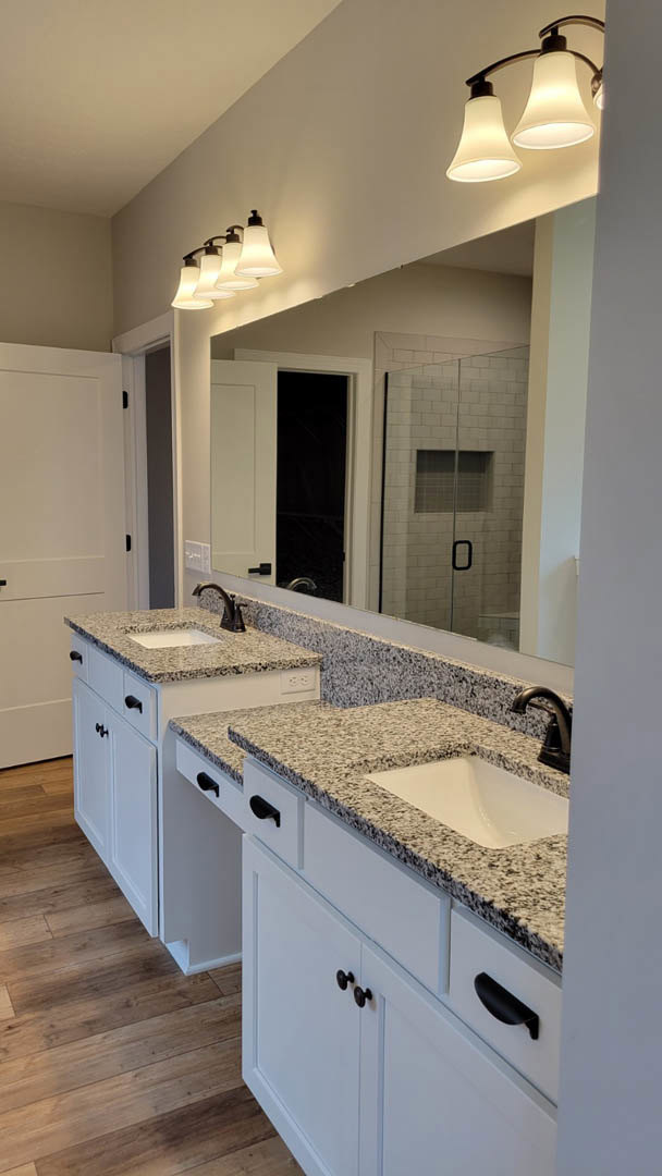 Bathroom with a wide framed mirror above dual white sinks set in black speckled countertops, modern light fixtures mounted on the wall, white cabinetry with black handles, glass
