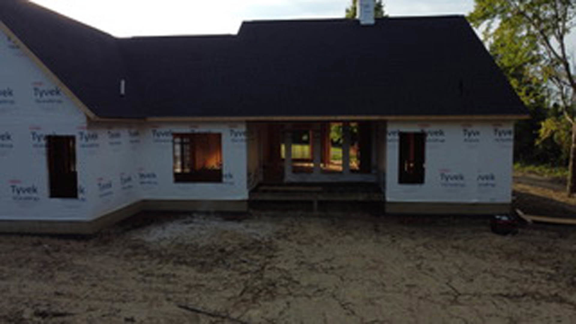 Partially built house with black roof, exposed framing, dirt yard, scattered construction materials, and multiple windows under a clear sky