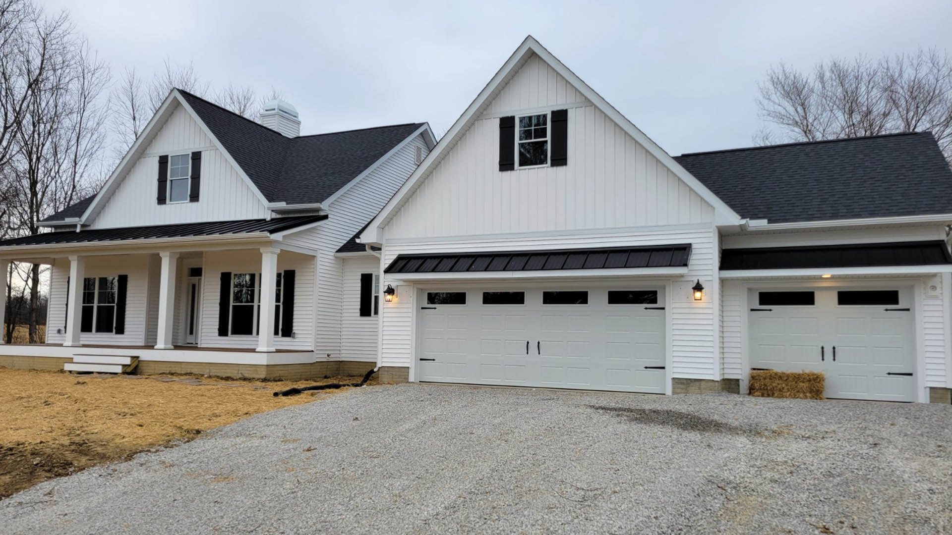 Two-story home with white siding, black shuttered windows, attached garage with white door, gravel driveway, and covered front porch under gabled roof