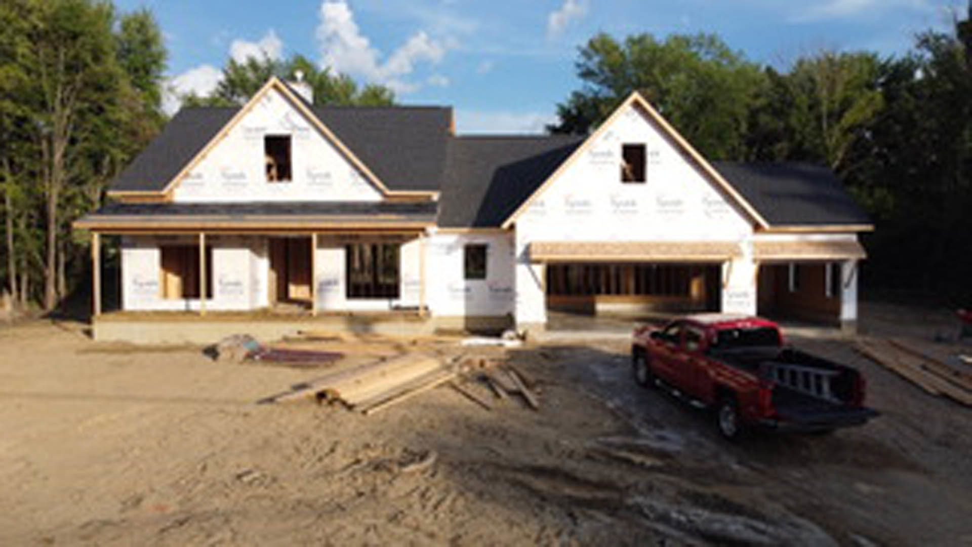 Red truck with ladder parked on dirt driveway in front of partially constructed house with exposed framing, blue sky overhead, trees in background