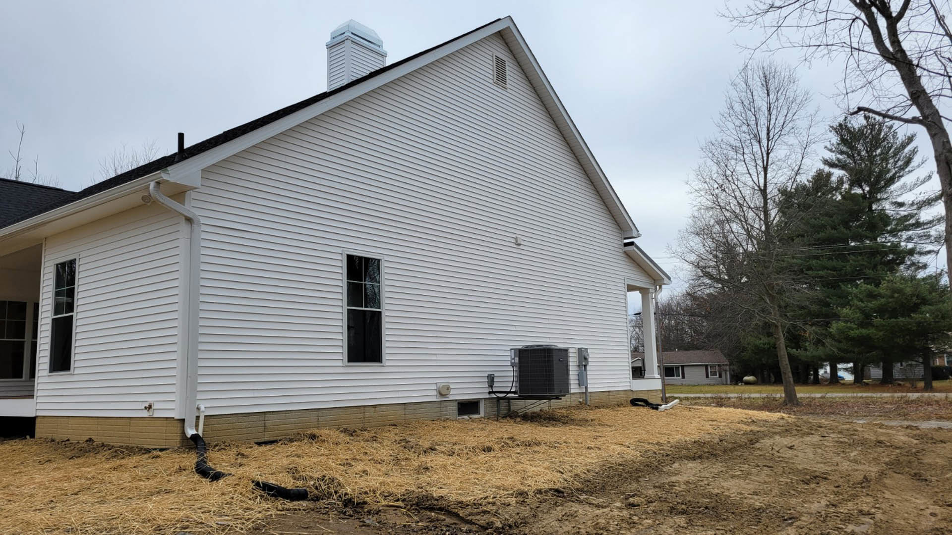 White house with horizontal siding, white-framed windows, and a white chimney on the roof, large grassy yard with patches of dirt, black HVAC unit mounted on exterior wall, mature