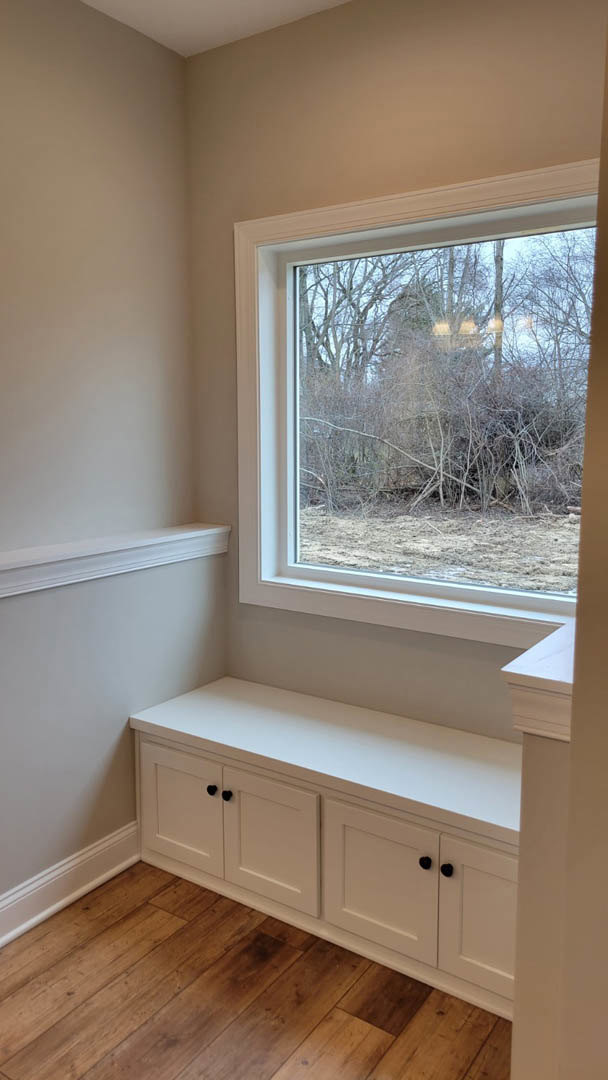 Large window with white blinds overlooks forest, wooden floor, white cabinets with black knobs, open cabinet door, white countertop, window seat bench along white wall