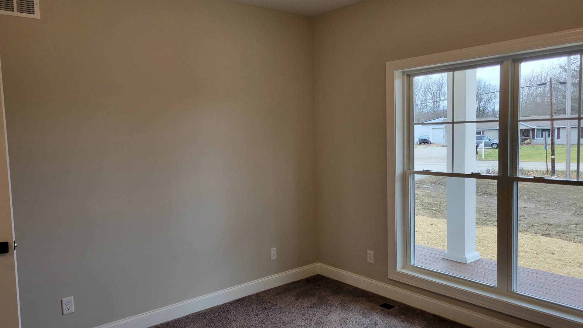 Carpeted room with a large window showing a dirt road and neighboring house, white wall outlet, door, and ceiling vent
