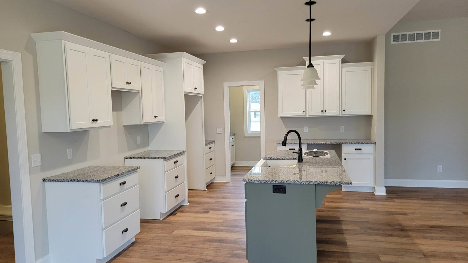 White kitchen with shaker cabinets, black handles, granite countertops, central island featuring built-in sink, stainless steel appliances, light wood flooring, and white door with