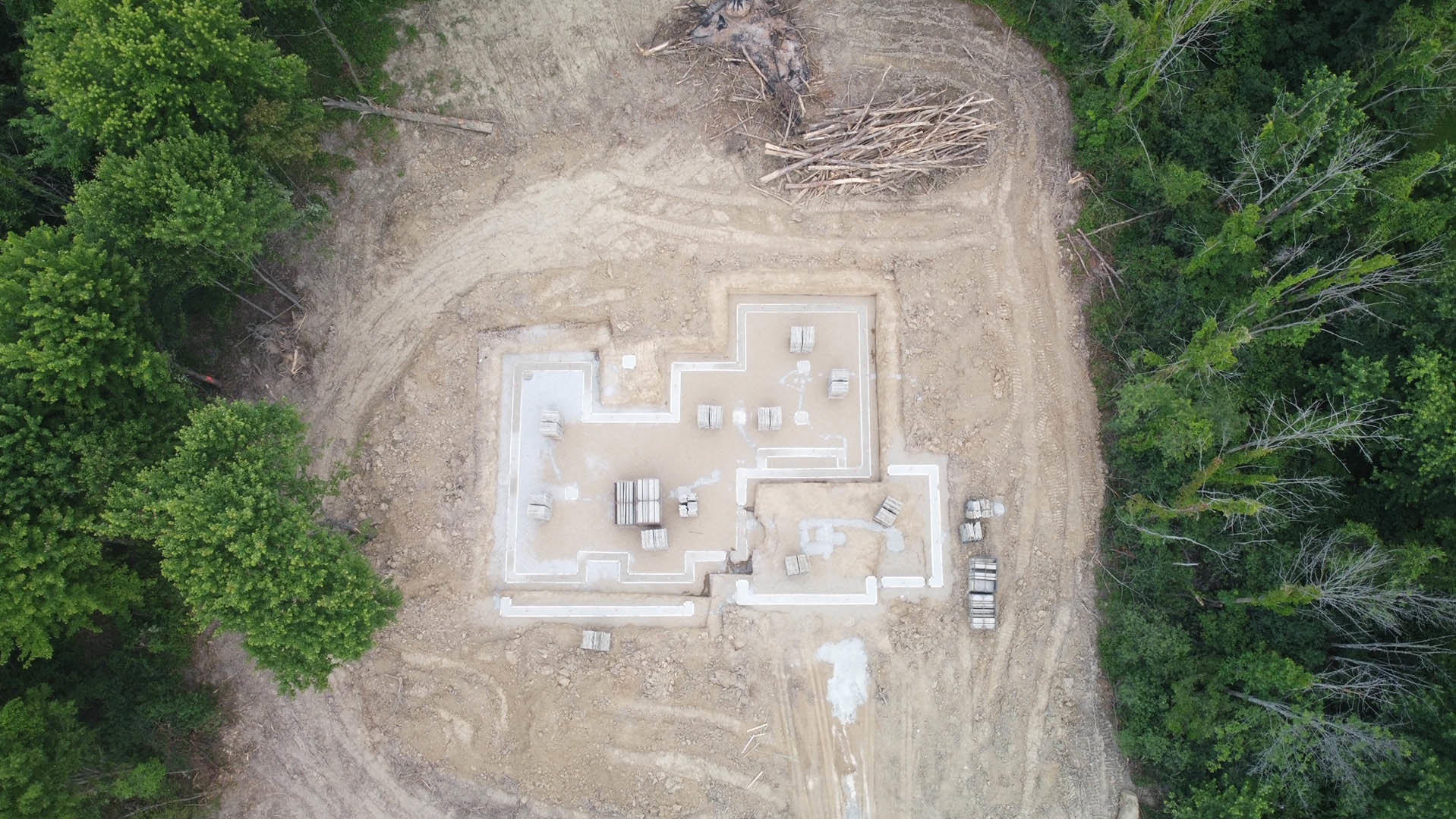 Square foundation set in dirt surrounded by trees, construction materials scattered nearby, dirt road leading to site