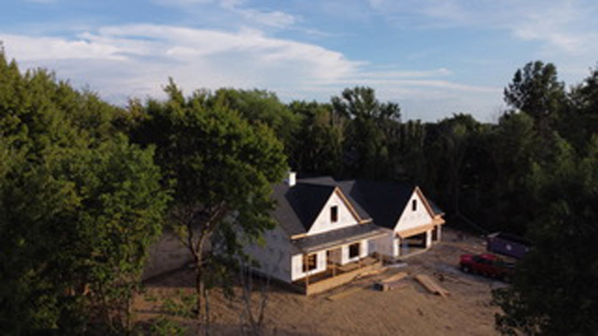 Framed custom home under construction with exposed wood beams, unfinished exterior walls, and leafy trees in the background