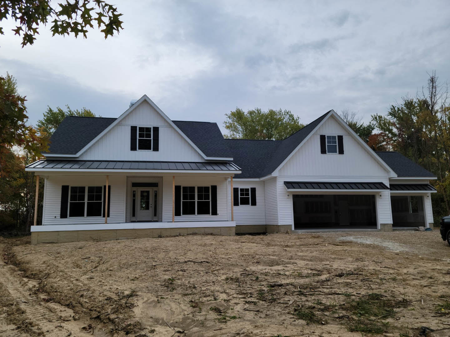 Black roof house with white siding, white-framed windows, and a white door, set beside a dirt yard and a tree in the background under a cloudy sky
