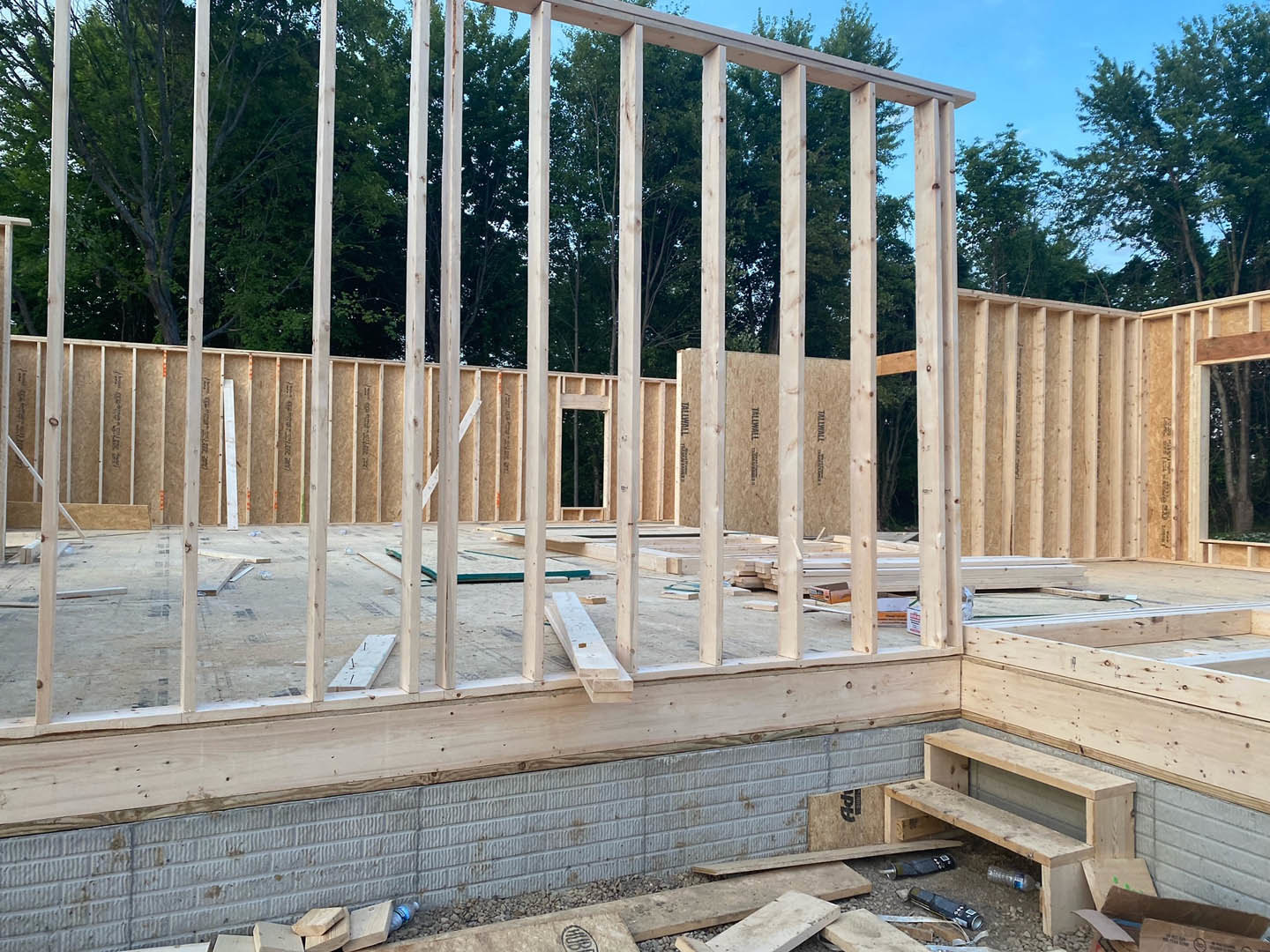 Wood framing and beams under construction atop a concrete foundation, with trees and blue sky in the background