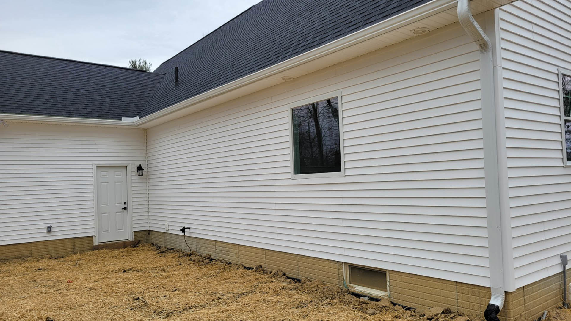 White siding exterior with a single white door featuring black handles, adjacent window reflecting trees, under a blue sky with scattered clouds.