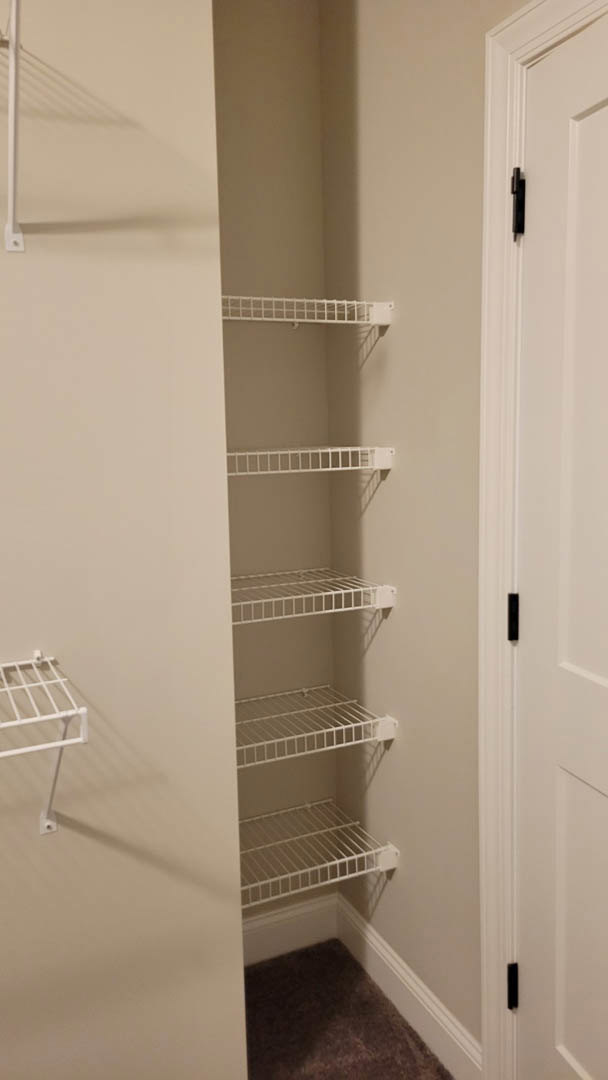 Walk-in closet with white shelving units, carpeted floor, white metal rack, and white walls, viewed near a partially open door