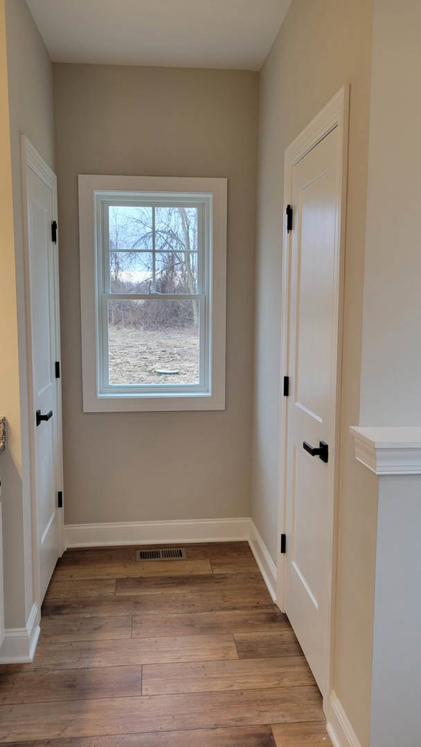 White interior room with wood laminate flooring, white walls, a tan accent wall, black door handles, window showing outdoor view, and floor vent near the baseboard.
