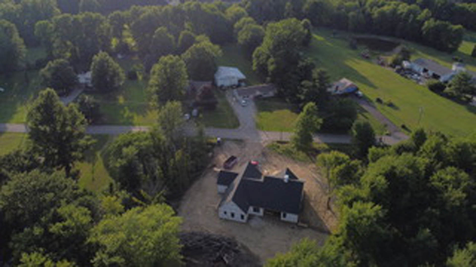 Two-story house with black roof and white siding, nestled among mature green trees, surrounded by grass and landscaped yard
