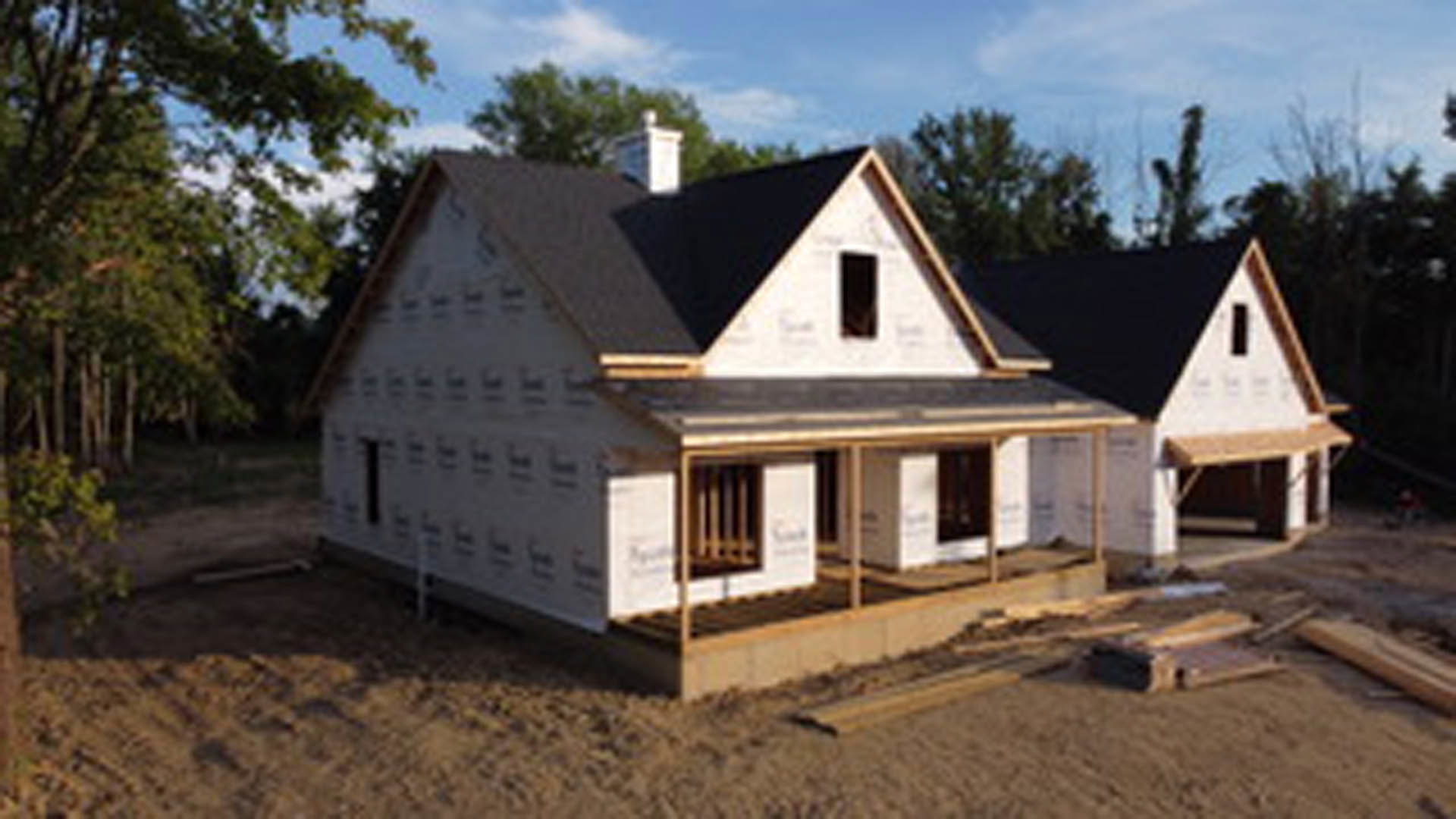 Two-story house under construction with exposed framing, partially finished roof, wooden deck, and several trees in the background