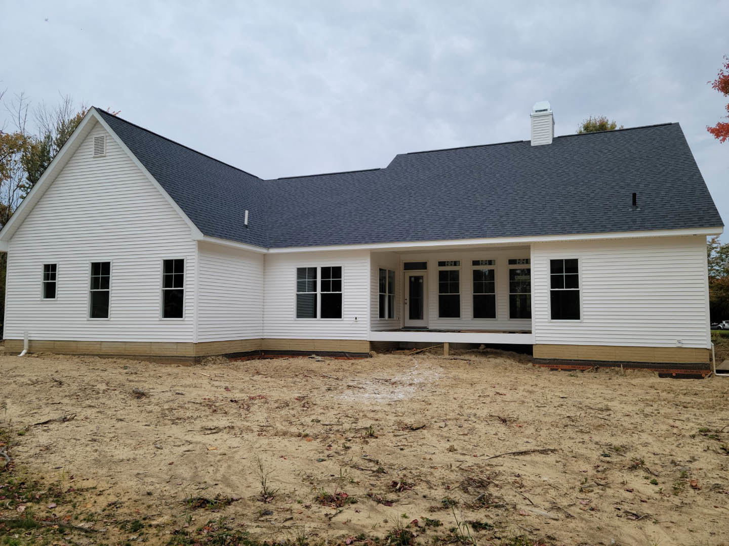 Partially built house with blue roof, white siding, and white-framed windows, surrounded by dirt ground and scattered plants under a cloudy sky