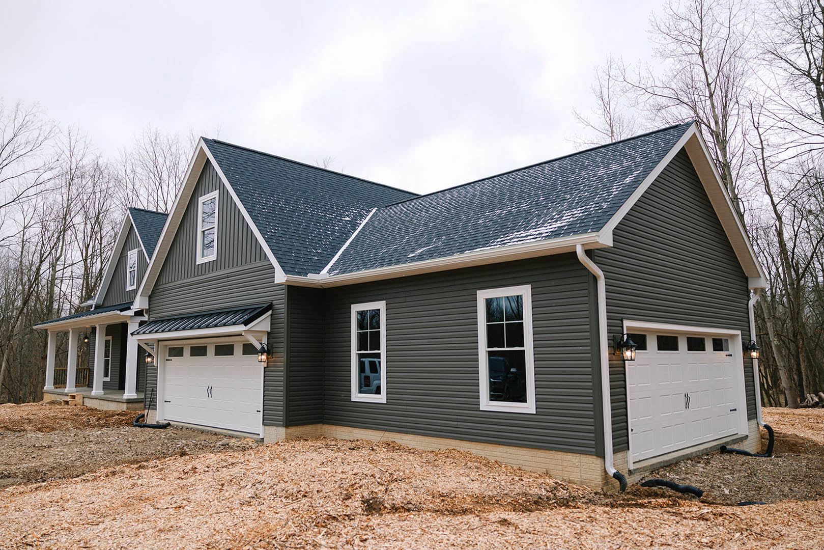 Two-story home with white siding, snow-covered roof, white-framed windows, attached garage with lantern, blue car visible through window, and pile of wood chips near driveway.