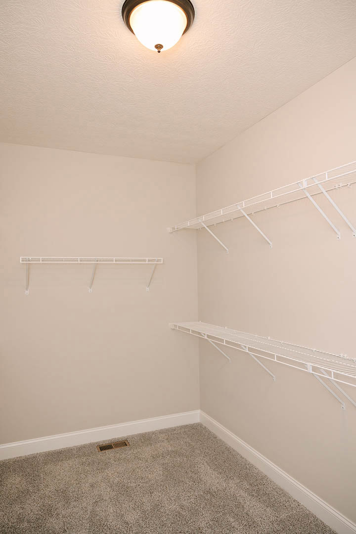Carpeted room with built-in white shelves, round ceiling light fixture, white metal railing, ceiling vent, exposed white pipe, and long white shelf against plaster walls.