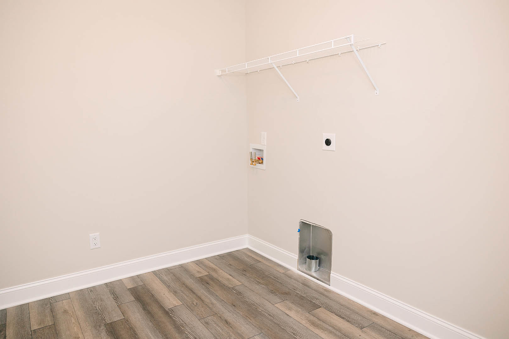Wood floor with white baseboards, white wall featuring a black round outlet, white metal shelf mounted above, silver container with black lid in the corner.