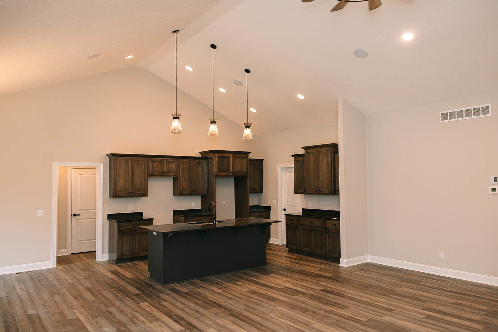 Dark wood kitchen cabinets, black island with matching countertop, white door featuring black handle, pendant light fixture, wall vent, slim metal pole, hardwood flooring