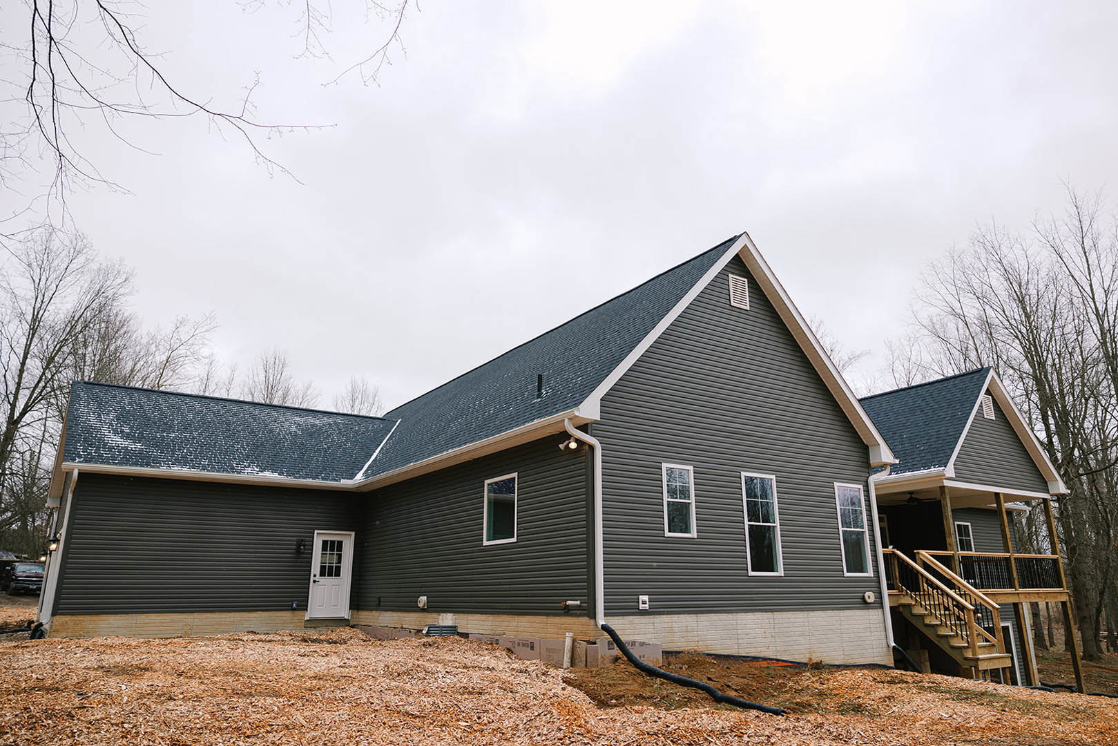 Two-story home with light siding, black-framed windows, white door with glass panes, and wooden staircase leading to entry, surrounded by trees and cloudy sky