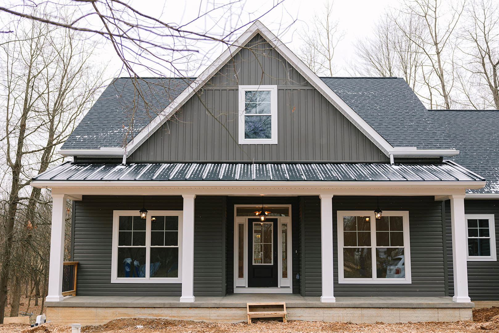 White cottage-style home with horizontal siding, black front door framed in white, covered porch with wooden steps, sash windows with white trim, exterior lighting, and mature tree