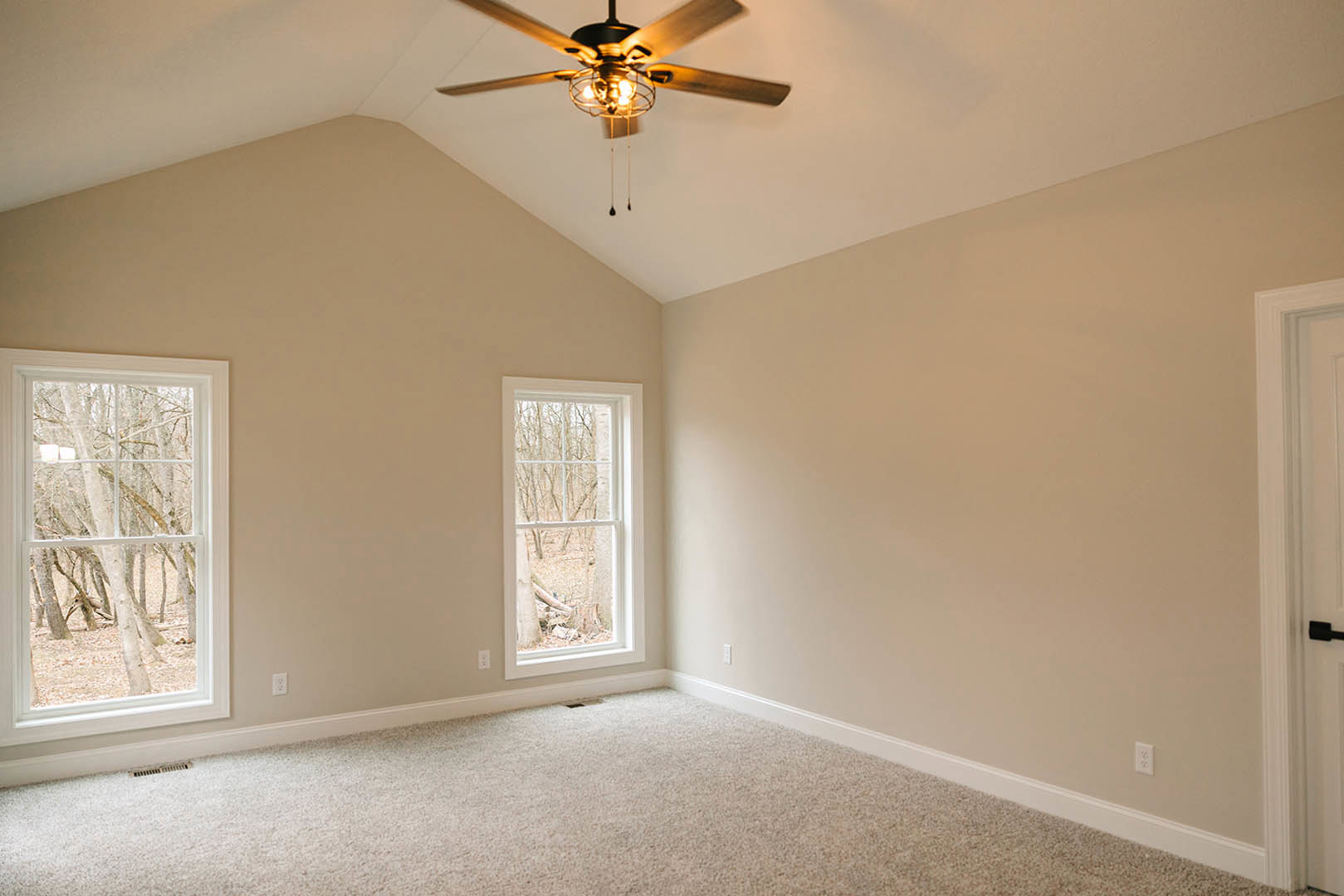 Ceiling fan with illuminated light fixture above carpeted floor, large window revealing leafy trees outside, white walls with crown molding