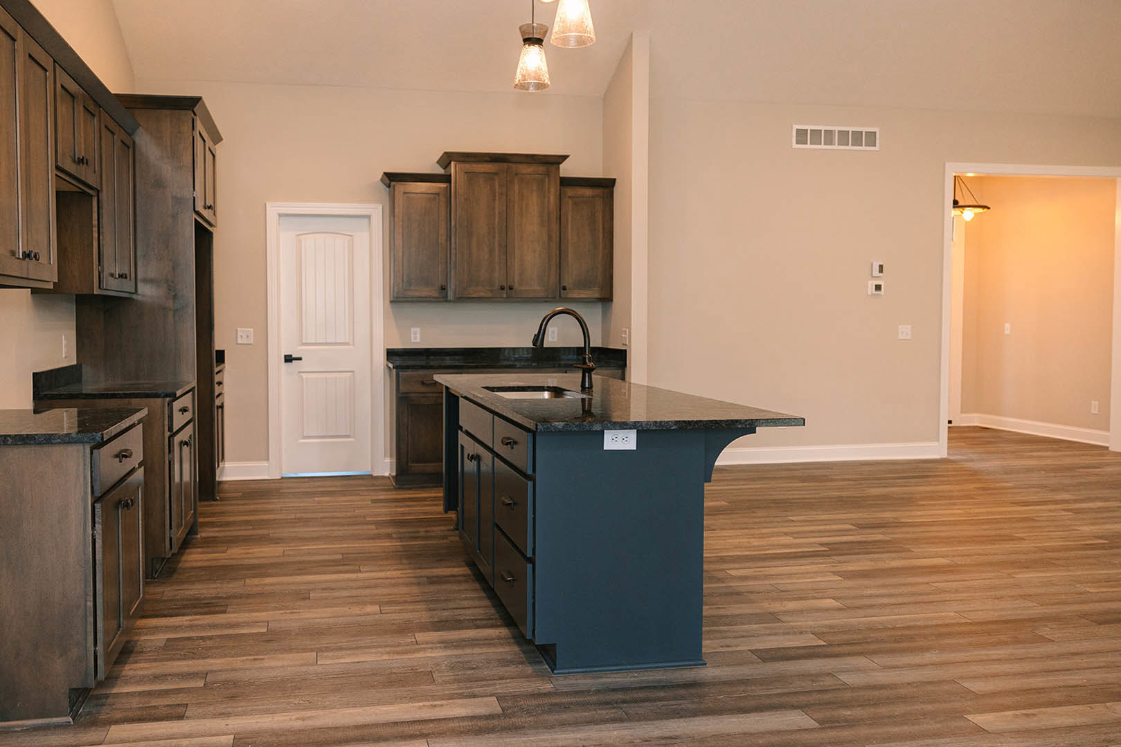 Kitchen with central island featuring built-in sink, wood flooring, row of wooden cabinets, white door with black handle, pendant light fixture, and close-up of lamp shade