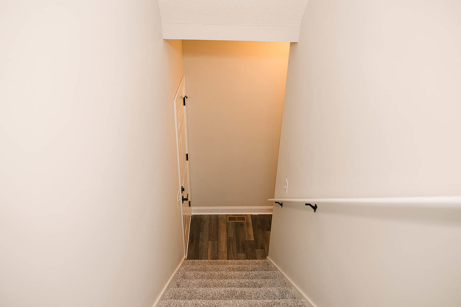 Wood staircase with carpet runner ascending toward a white door, wood flooring with vent, white plaster walls, ceiling light fixture, black wall brackets, and tile bathroom visible