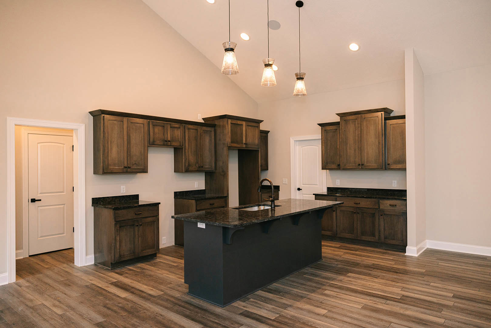 Black kitchen island with built-in sink, wooden cabinets with black countertops, white door with black handle, pendant lighting, hardwood flooring