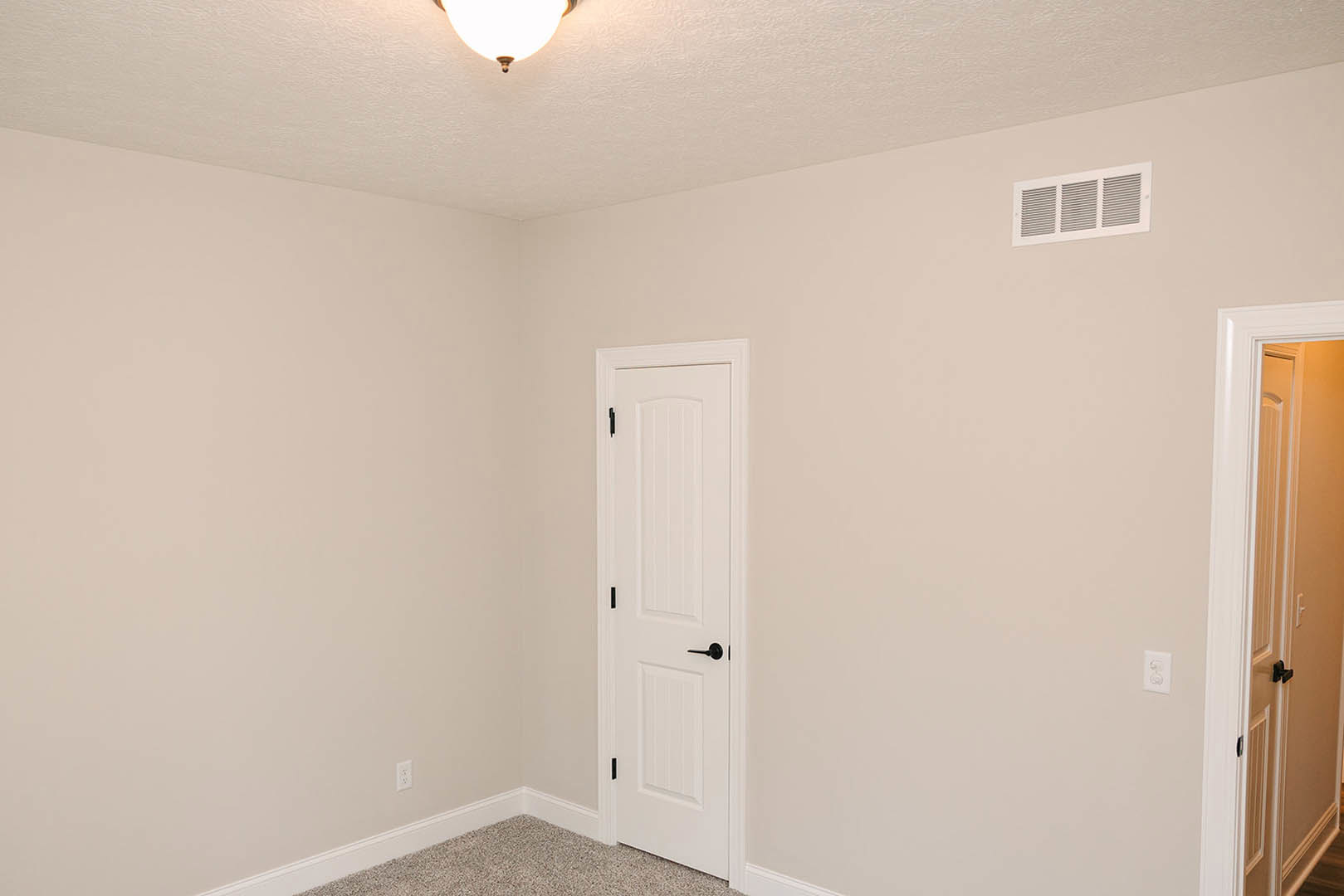 White paneled door with black handle, white plaster walls, vent above baseboard, round ceiling light fixture, light wood flooring, decorative wall molding