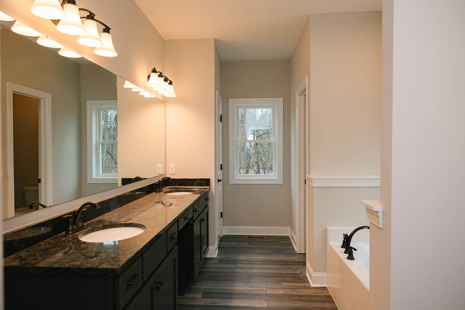 Bathroom featuring marble countertop with undermount sink, freestanding tub near window overlooking trees, wood flooring with vent, modern light fixture, and large mirror above