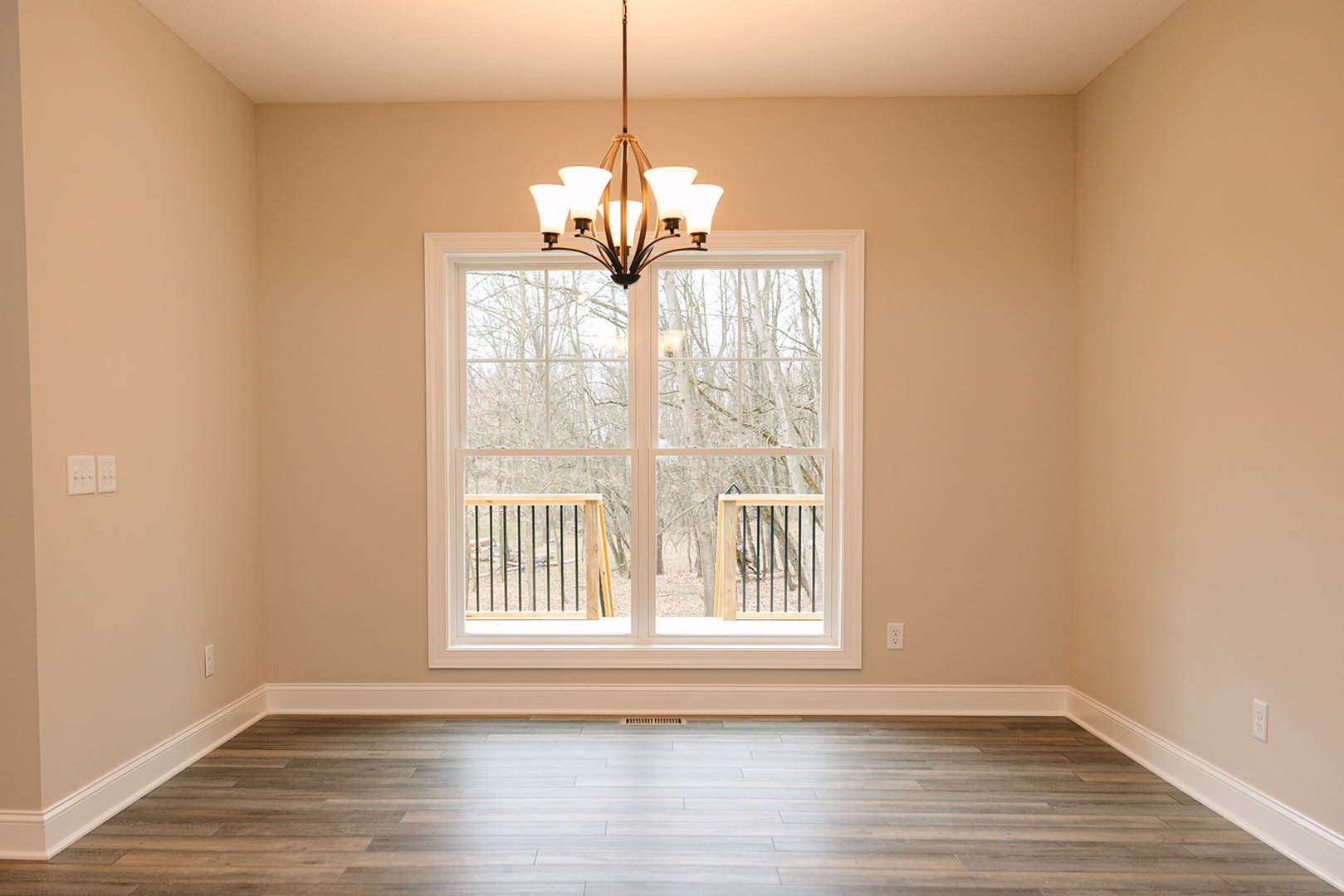 Bright room with large window overlooking trees, white wood flooring with vent, wooden railing featuring black metal bars, ornate chandelier hanging from plaster ceiling, soft