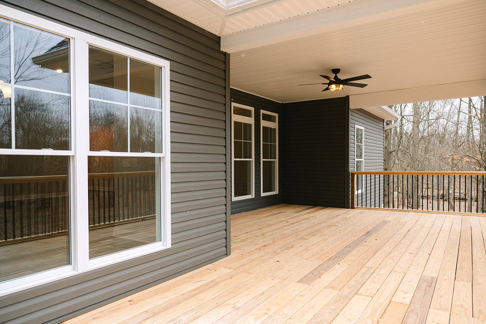 Covered deck with wooden flooring, white-framed windows, grey walls, ceiling fan with light, and metal railing
