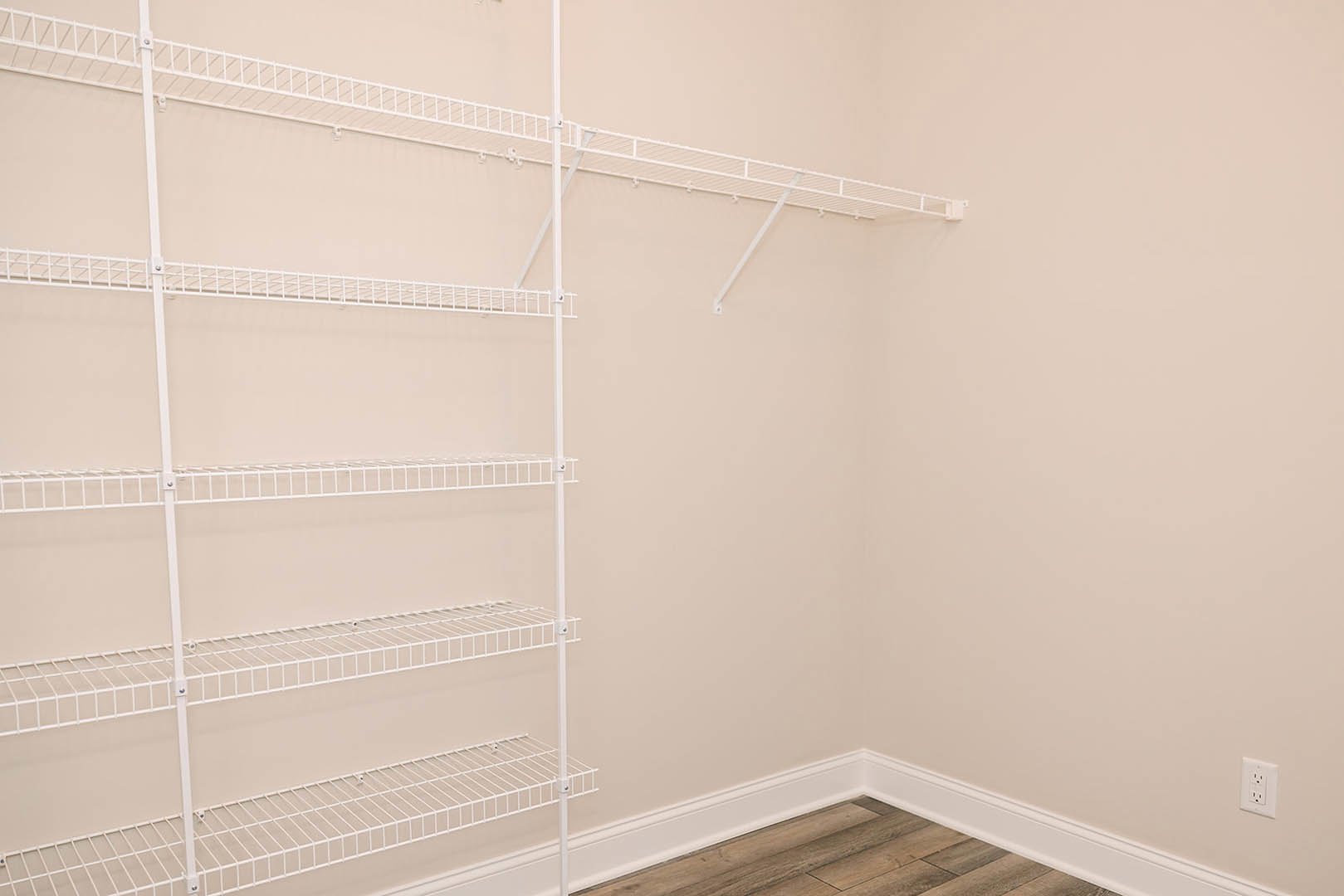 White wire shelving unit with six bars mounted on a white plaster wall above white baseboard, close-up view includes electrical outlet.