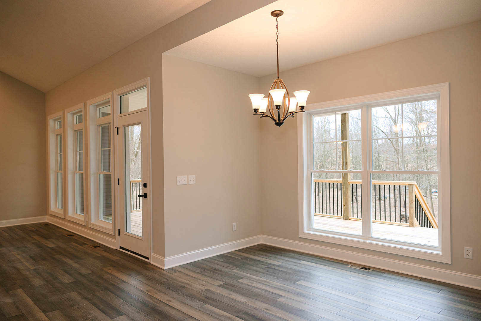 Chandelier hanging from a white ceiling above wood flooring, large windows with molding framing a view of a balcony with railing, neutral walls and decorative trim