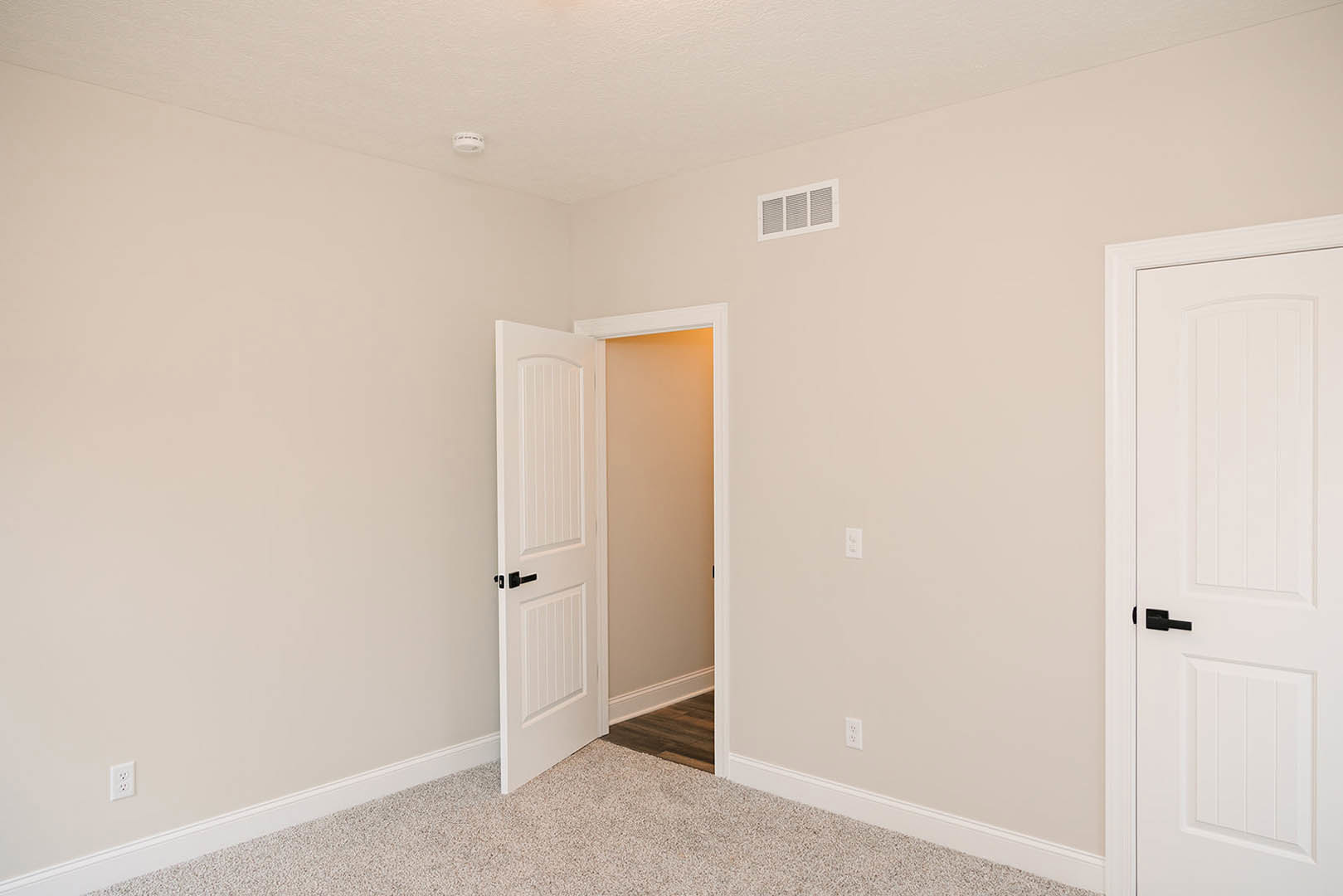 White paneled door with black handle opening into carpeted room, white walls with wall vent and sconce visible