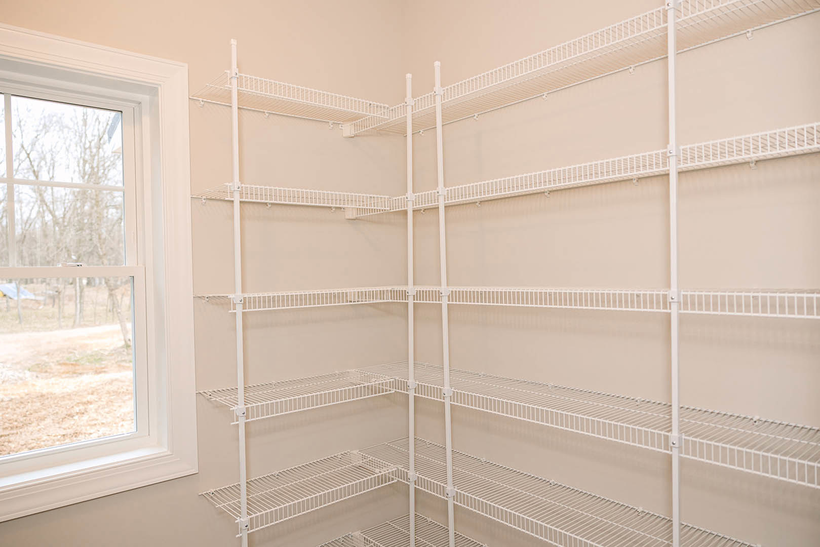 White metal shelving unit with attached ladder beside a large window overlooking leafy trees, set against a neutral wall in a modern room.