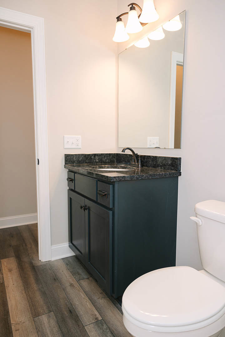 Bathroom with white tile walls, wood flooring, modern sink and faucet, rectangular mirror above countertop, close-up of toilet and light fixture, white wall outlet with knobs.