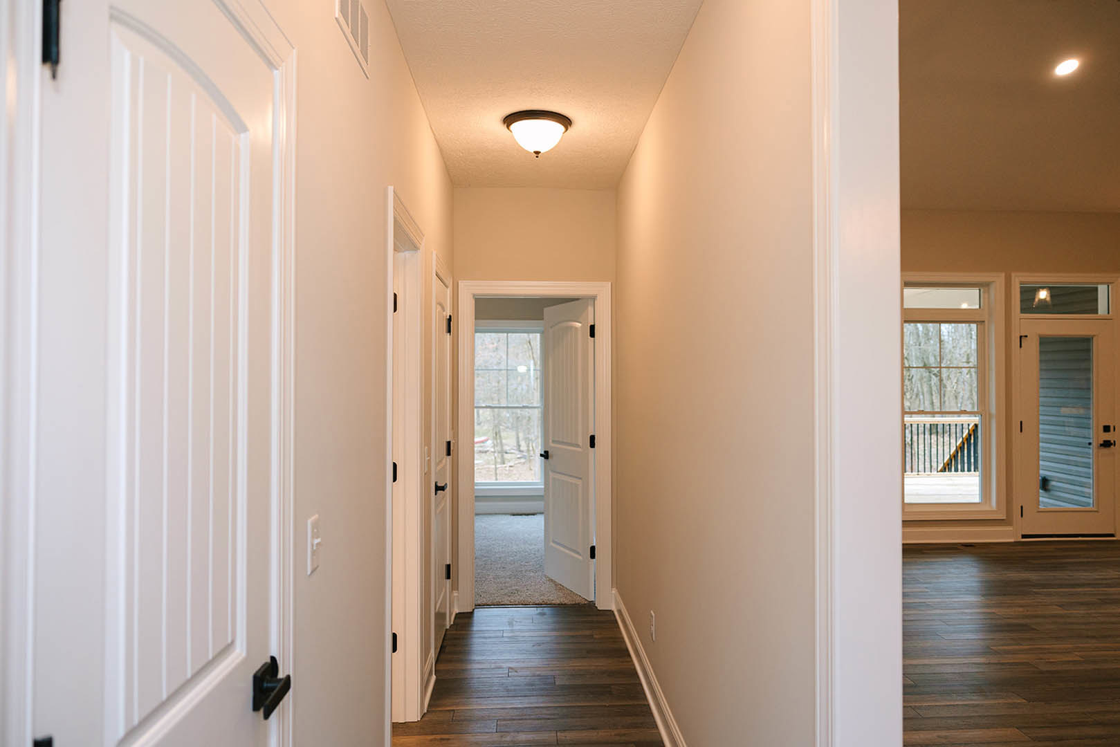 Hallway with dark hardwood floor, white walls and doors, ceiling light fixture, window with railing offering outdoor view, sunlight reflecting on wood surface