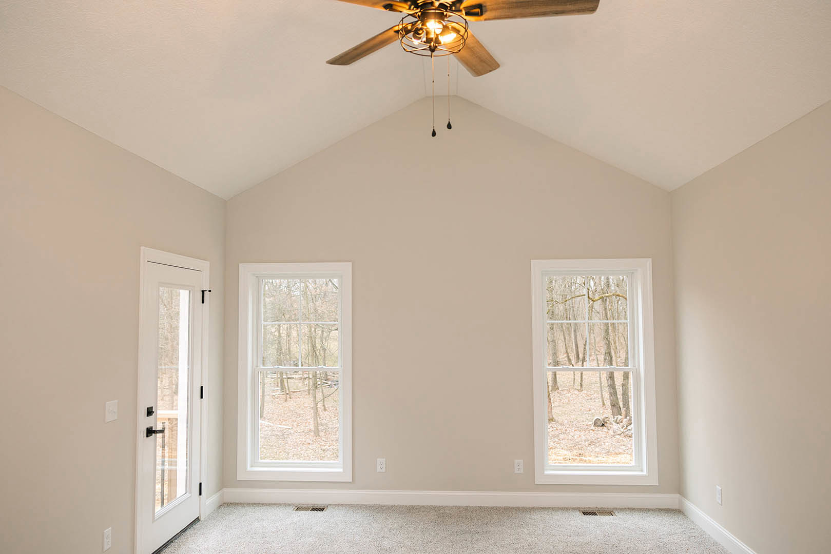 Ceiling fan with illuminated light fixture mounted on white ceiling, carpeted floor, white walls, window showing trees outside, door with handle and doorknob