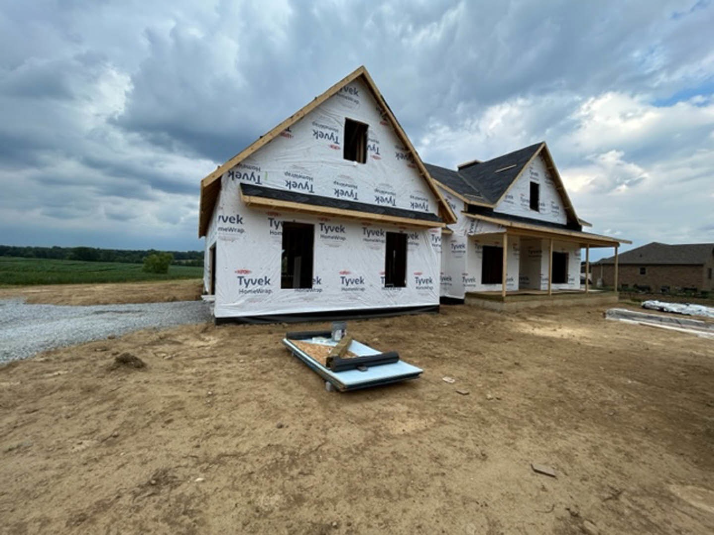 Framed house under construction with exposed wood, white plastic sheeting, and open windows, set beside a grassy field under a cloudy sky