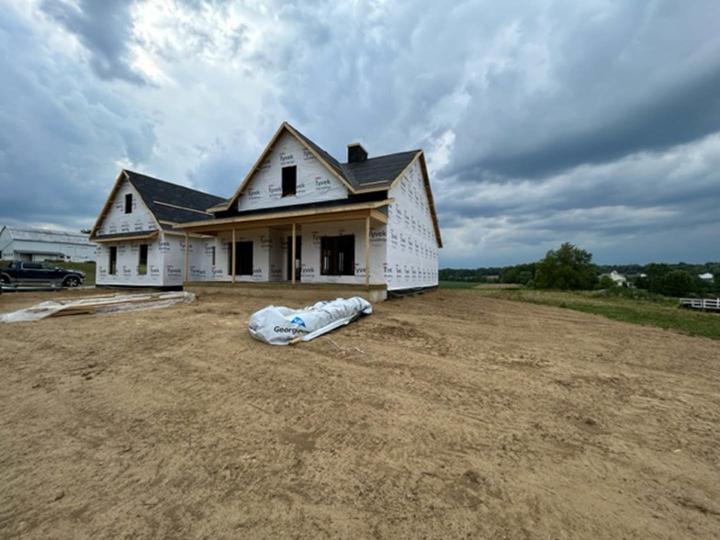Partially built house with exposed framing and roof, white construction bag on grassy ground, cloudy sky overhead