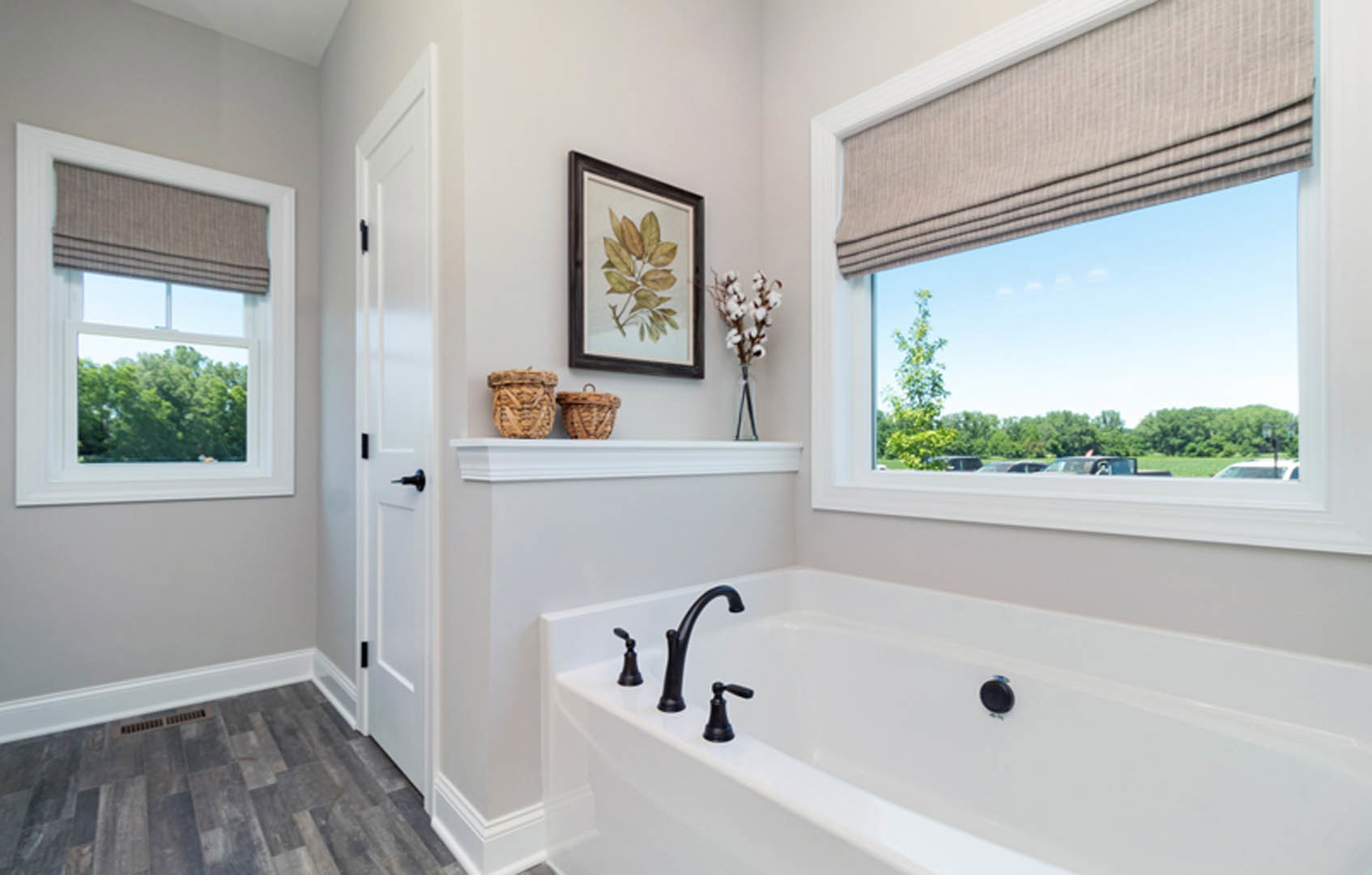 Freestanding white bathtub beneath a large window with a light-filtering shade, neutral tile flooring, chrome faucet, and potted green plant on the sill