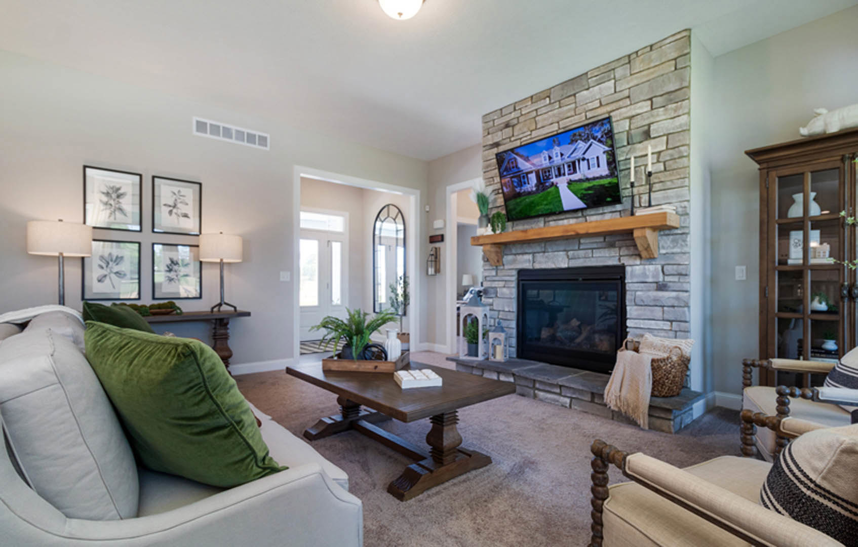 Living room with a television mounted above a glass-door fireplace, green pillow on a couch, wooden coffee table with a white decorative object, brick wall displaying a framed
