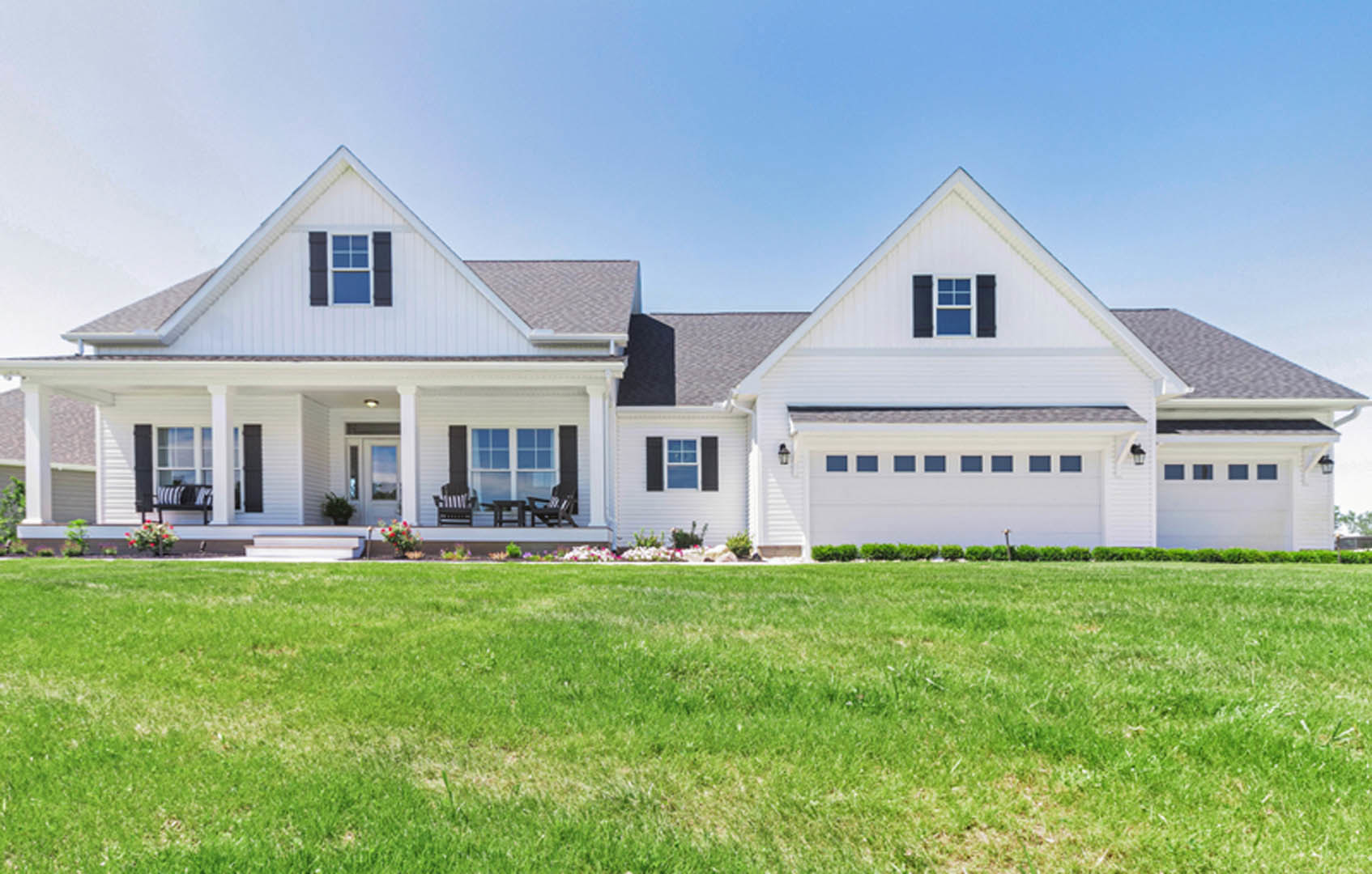 White farmhouse-style home with white framed windows, covered porch, attached garage, and manicured green lawn under a clear sky