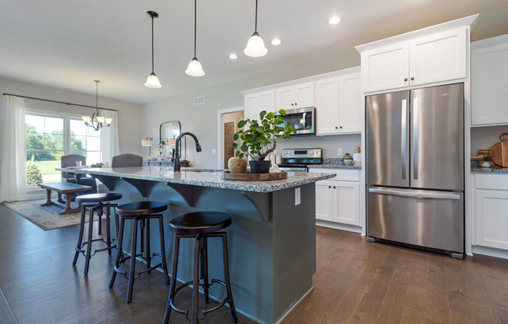 Modern kitchen featuring a marble-topped bar with round wooden stools, stainless steel refrigerator, white cabinetry, potted plant on the counter, and light hardwood flooring