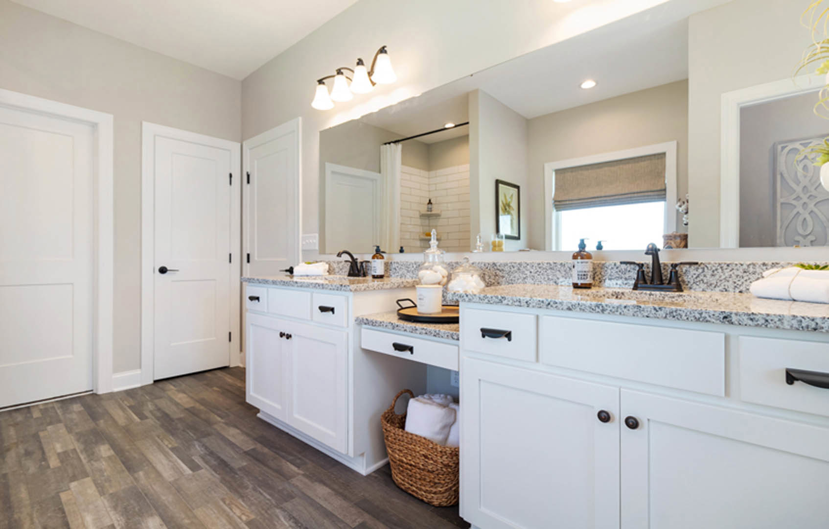 Bathroom with white cabinets, marble countertops, basket with white pillow, row of light fixtures, white candle on table, and blurred plant in background