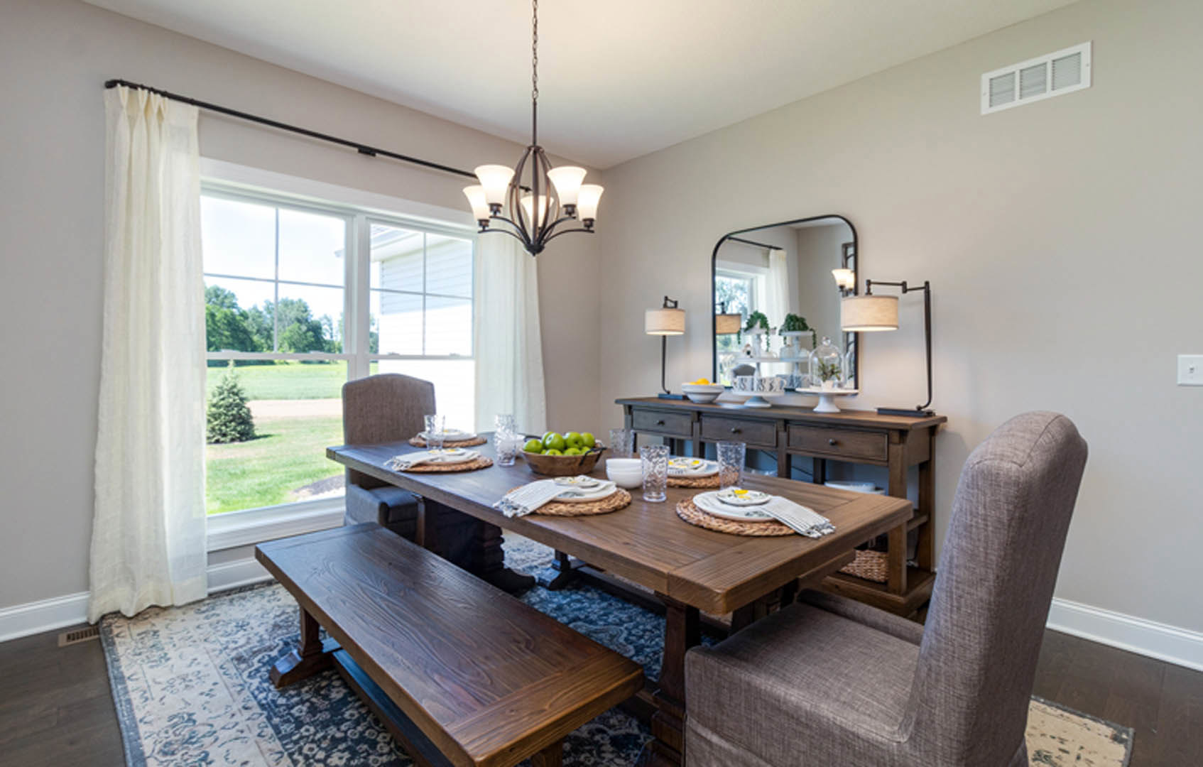 Dining room with wooden table set with plates and glasses, matching bench on patterned rug, large wall mirror, bowl of green apples, sheer white curtains, and neutral finishes