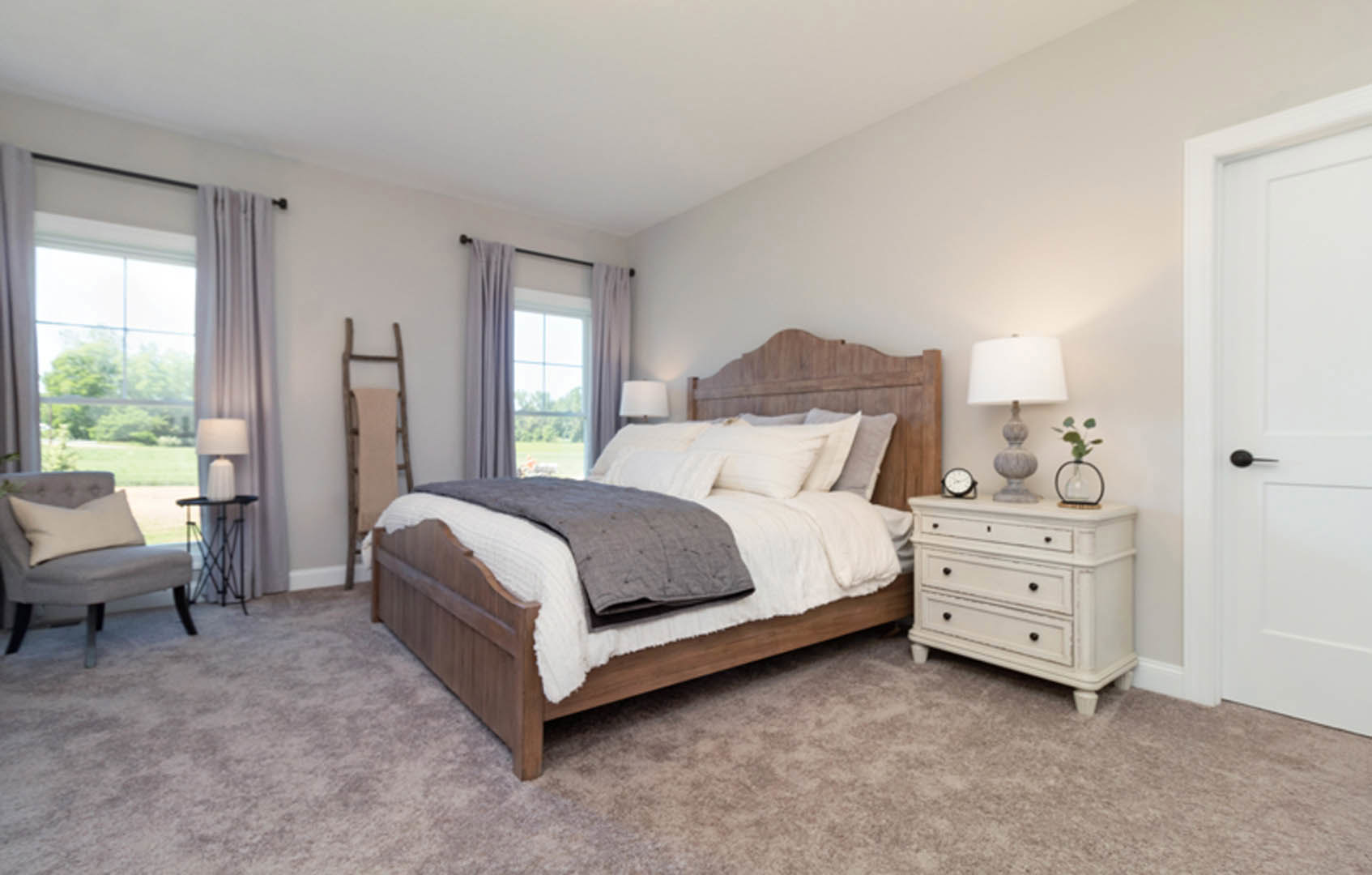Bedroom with a wooden headboard bed, white dresser with black knobs, nightstand holding a lamp, upholstered chair with pillow, neutral walls and flooring