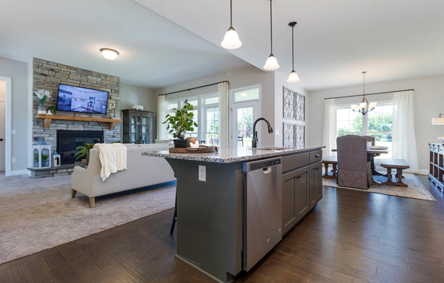 Open-concept kitchen and living room featuring white cabinetry, stone countertops, television mounted on a brick accent wall above a fireplace, potted plant, dining table with