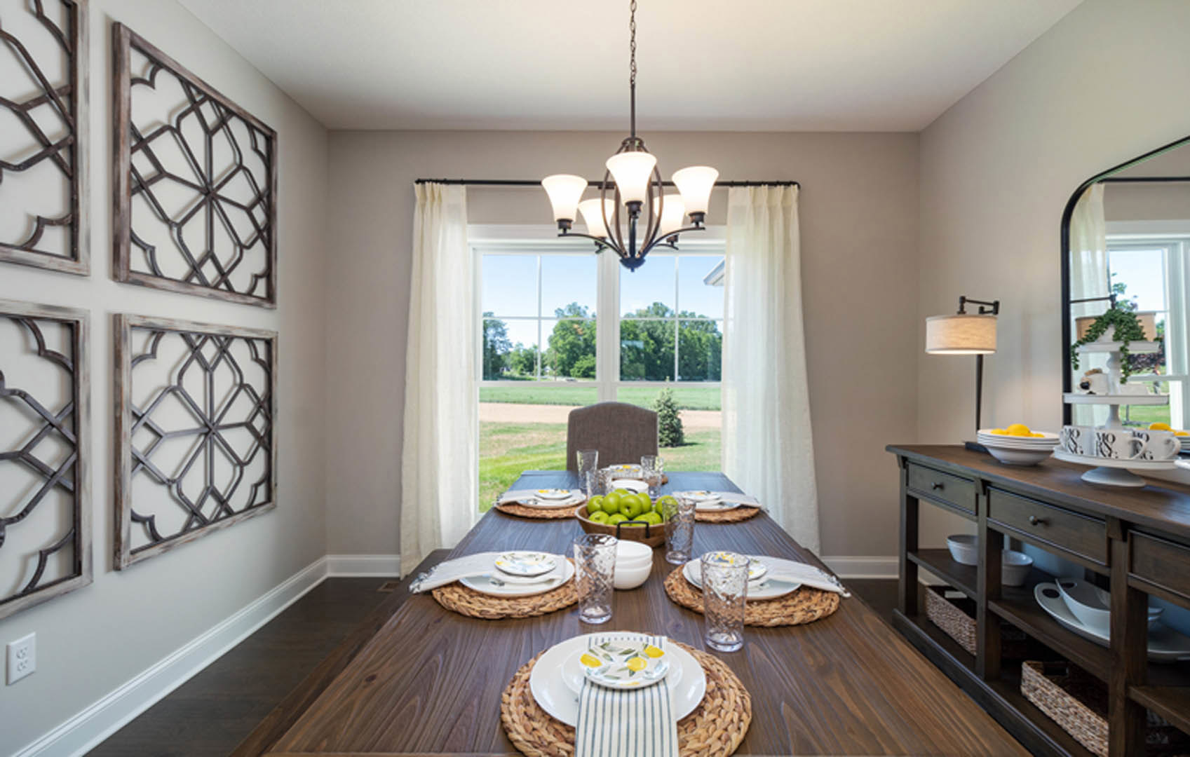 Wood dining table set with plates, napkins, glasses, and a bowl of fruit, surrounded by upholstered chairs in a bright dining room with cabinetry, windows, and decorative vases.
